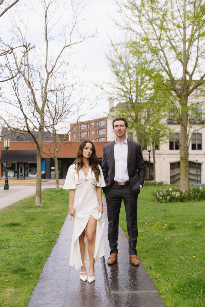 A woman in a white dress and heels stands next to a man in a dark suit and brown shoes on a park path, surrounded by green grass and budding trees, with buildings in the background.