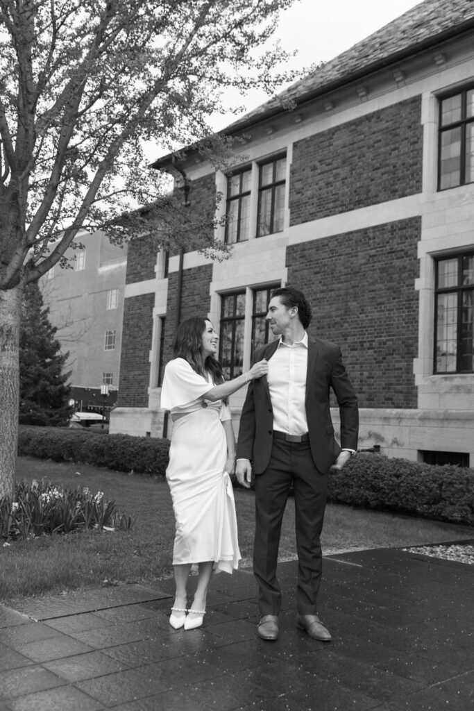 A woman in a white dress playfully adjusts the tie of a man in a suit as they stand together on a sidewalk outside a brick building, surrounded by trees and bushes.