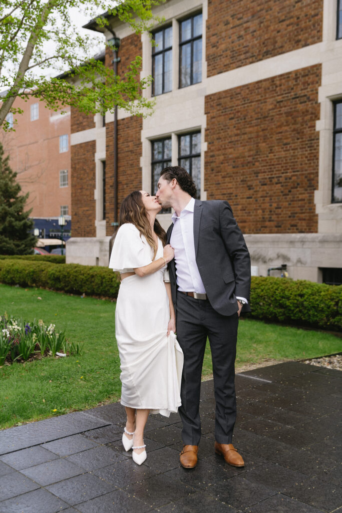 A couple dressed in wedding attire share a kiss outdoors in front of a brick building, with greenery and flowers around them. The woman wears a white dress and heels, and the man wears a dark suit with brown shoes.