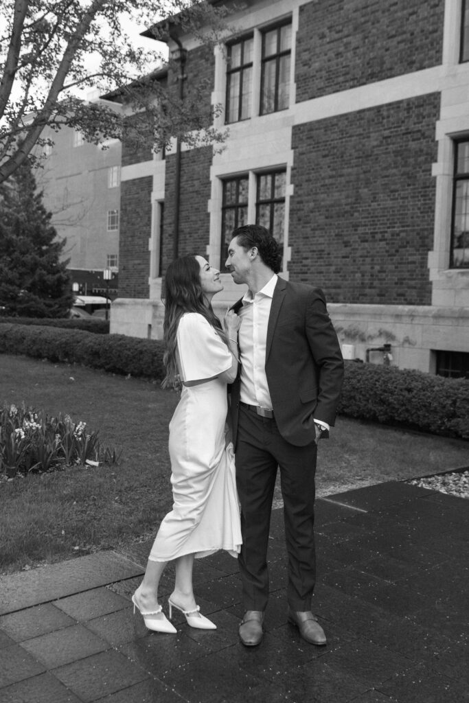 A couple dressed formally shares a joyful, intimate moment on a sidewalk outside a brick building, smiling at each other. The scene is in black and white, with greenery and flowers nearby.