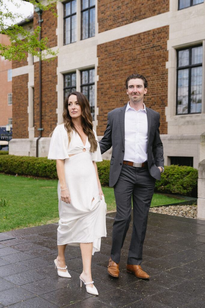 A woman in a white dress and heels walks beside a man in a gray suit and brown shoes on a wet sidewalk in front of a brick building with large windows.