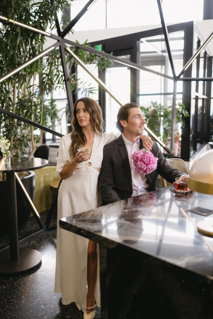 A woman in a white dress stands beside a seated man in a suit with pink flowers on his lap. They are in a modern, stylish lounge with plants and geometric decor; both have drinks and a relaxed, elegant demeanor.
