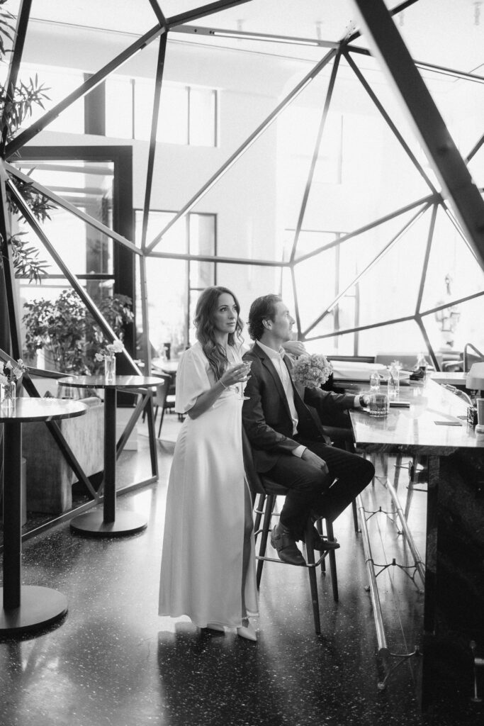 A woman in a long dress stands beside a seated man in a suit at a modern, geometric bar. Sunlight streams through large windows, creating a bright, airy atmosphere. Both appear relaxed and engaged.