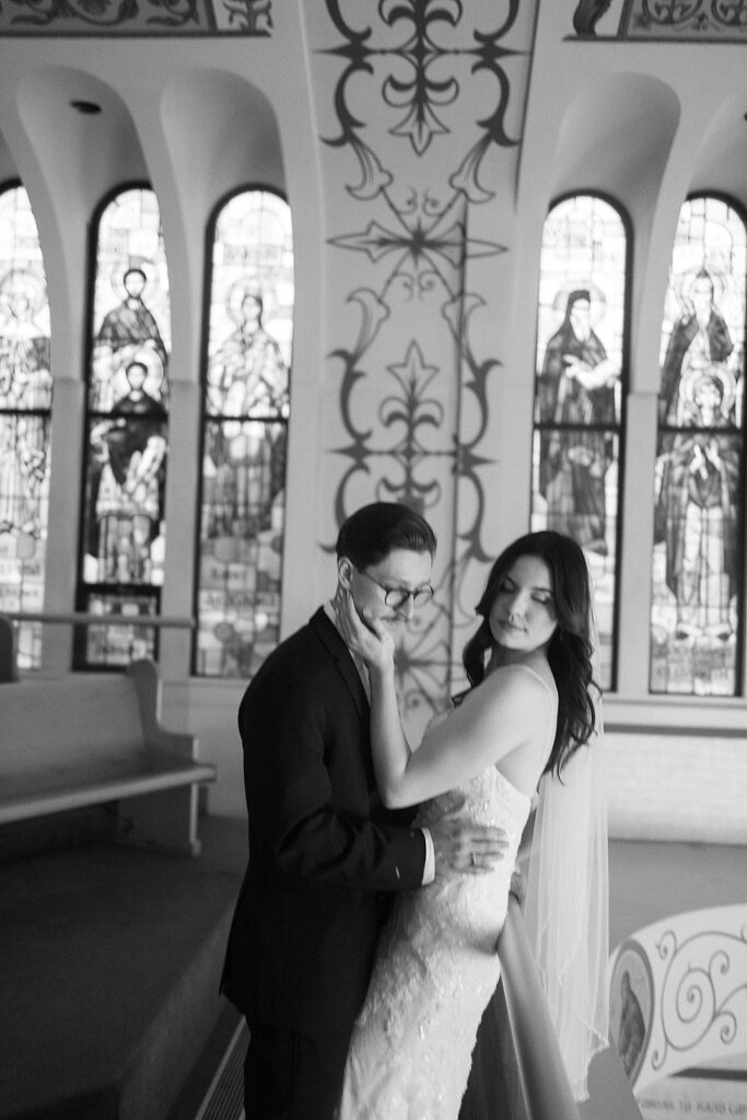 A black-and-white photo of a couple in formal attire embracing inside a church with tall, ornate stained glass windows in the background. The scene appears romantic and intimate.