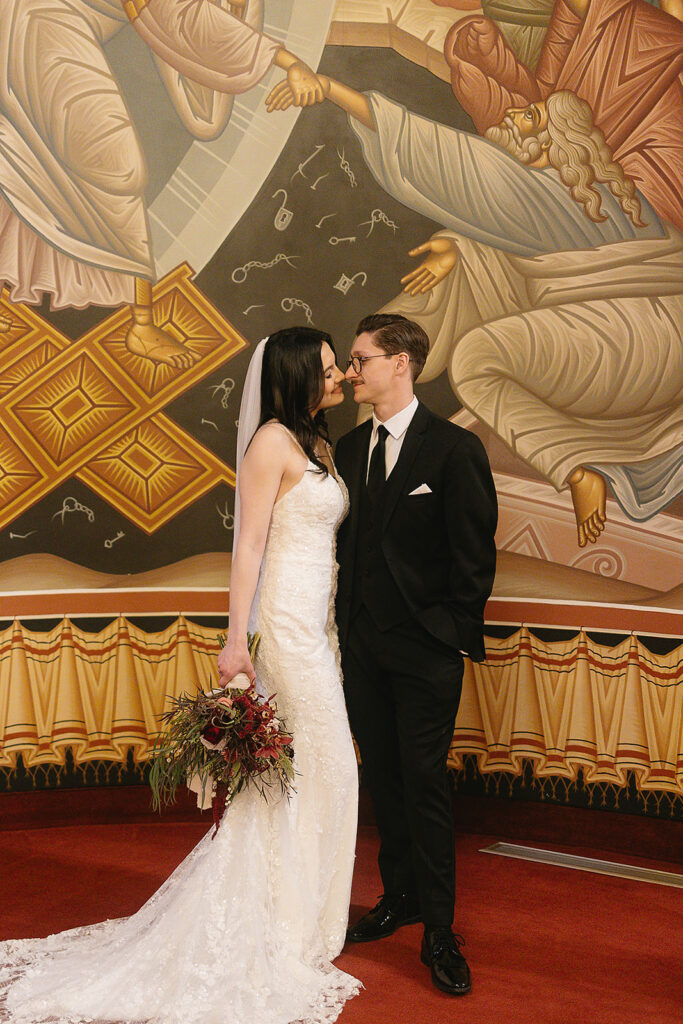 A bride in a white gown and veil stands close to a groom in a black suit. They face each other, smiling, in front of a colorful religious mural. The bride holds a bouquet of flowers.