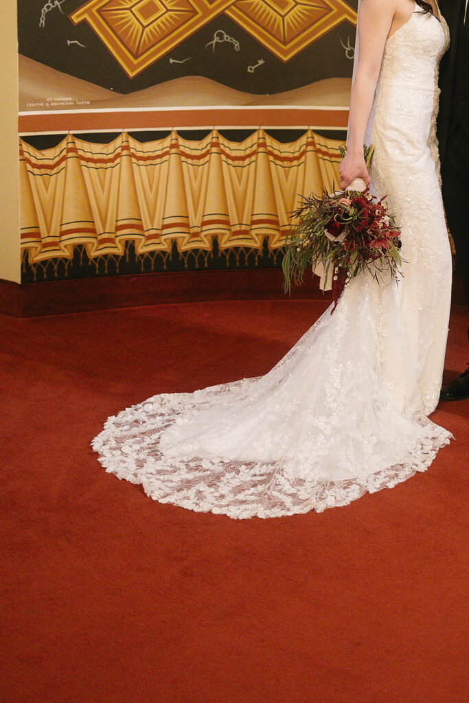 A bride in a white lace wedding gown holds a bouquet of dark flowers, standing on red carpet with ornate gold and tan wall decor in the background. The groom is partially visible beside her.