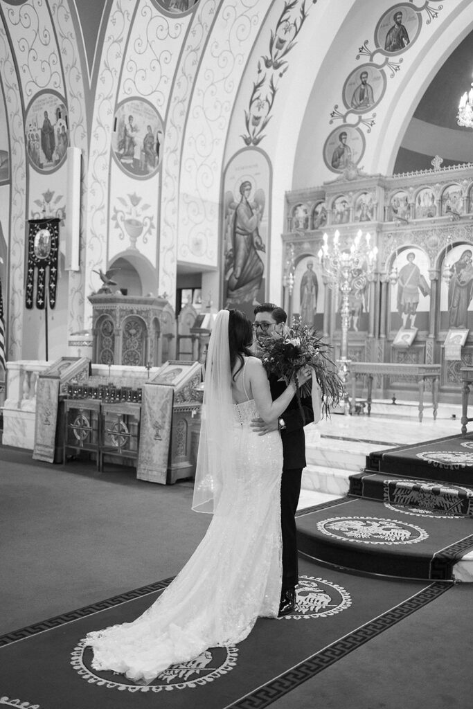 A bride and groom embrace at the altar of an ornately decorated church with religious icons and detailed frescoes on the walls, beneath a large chandelier. The bride wears a long veil and fitted gown.