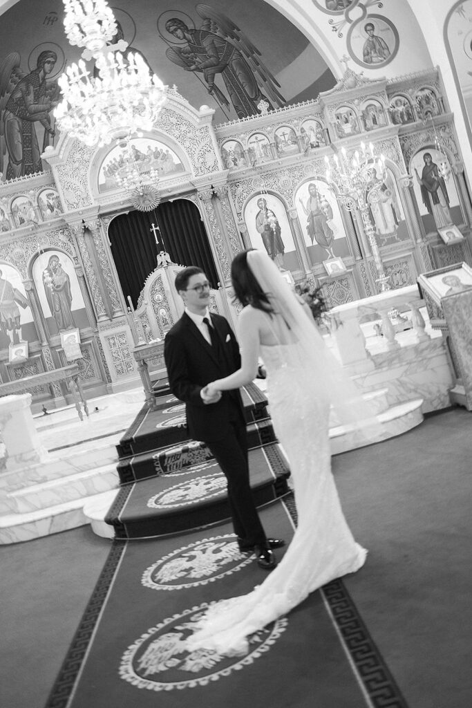 A bride and groom hold hands and face each other inside an ornate church, decorated with religious icons and chandeliers. The bride wears a veil and gown, and the groom is in a suit. The image is in black and white.