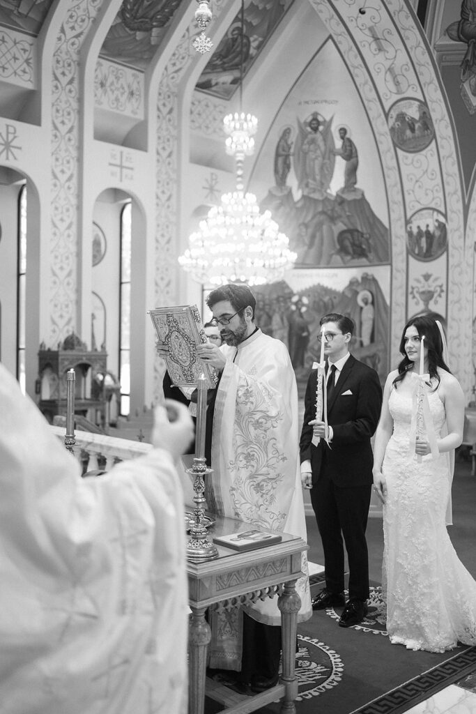 A priest conducts a wedding ceremony in an ornately decorated church, holding a book. A bride in a white dress and a groom in a dark suit stand side by side holding candles. The scene is black and white.