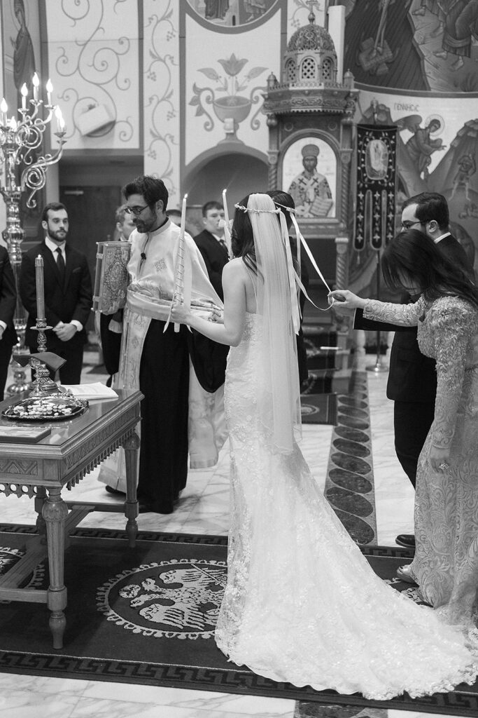 A bride and groom stand at the altar in a church during a wedding ceremony. The bride wears a long white gown and veil, holding candles, while attendants and a priest participate in the ritual. The scene is ornate and formal.
