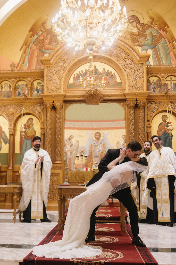 A bride and groom share a dramatic kiss at the altar of an ornate church, surrounded by priests in white robes and golden religious icons, under a grand chandelier.