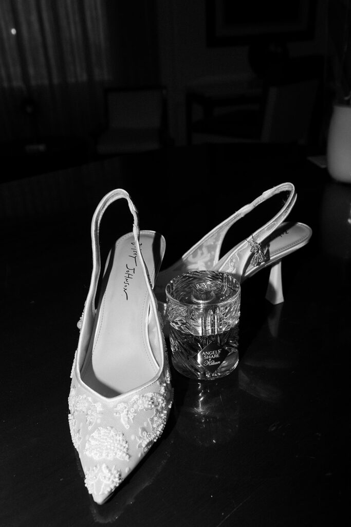 A pair of elegant, beaded slingback high-heeled shoes and a bottle of Chanel perfume sit on a dark surface in a softly lit room.