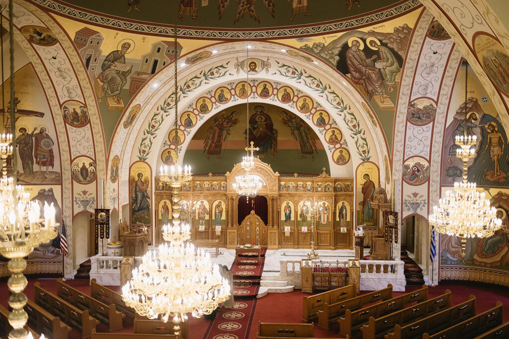The interior of an ornate Orthodox church featuring gold chandeliers, red carpet aisles, decorative arches, religious icons, and detailed murals covering the walls and ceiling above the wooden altar area.