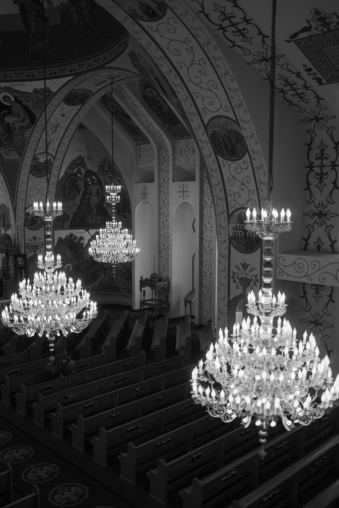 Black and white photo of a church interior with ornate chandeliers hanging above empty pews; religious murals and intricate wall designs are visible on the arched ceilings and walls.
