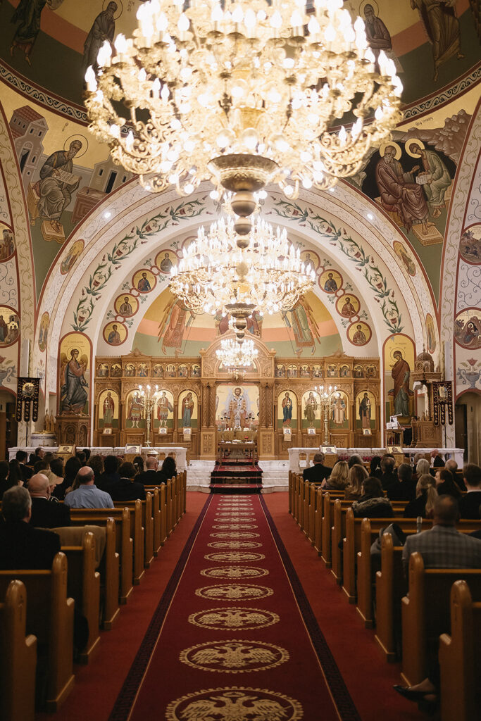 A grand, ornate church interior with golden chandeliers, religious icons, and intricate wall paintings. People sit in pews facing an altar at the end of a red carpeted aisle.