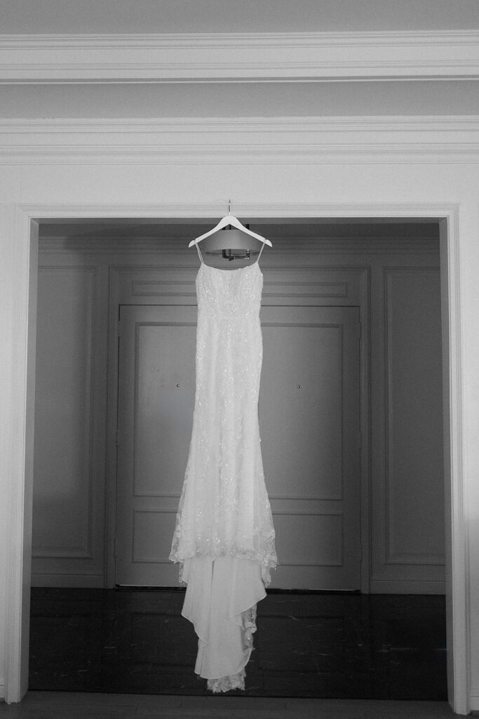 A white, sleeveless wedding dress hangs on a hanger in a doorway, with delicate lace details, set against a neutral interior background.