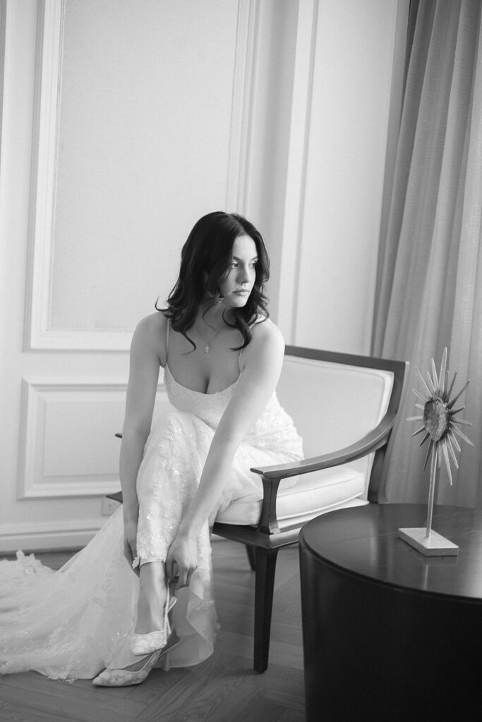 A woman in a white dress sits on a chair, looking to the side as she adjusts her shoe. The scene is indoors with elegant decor and soft light.