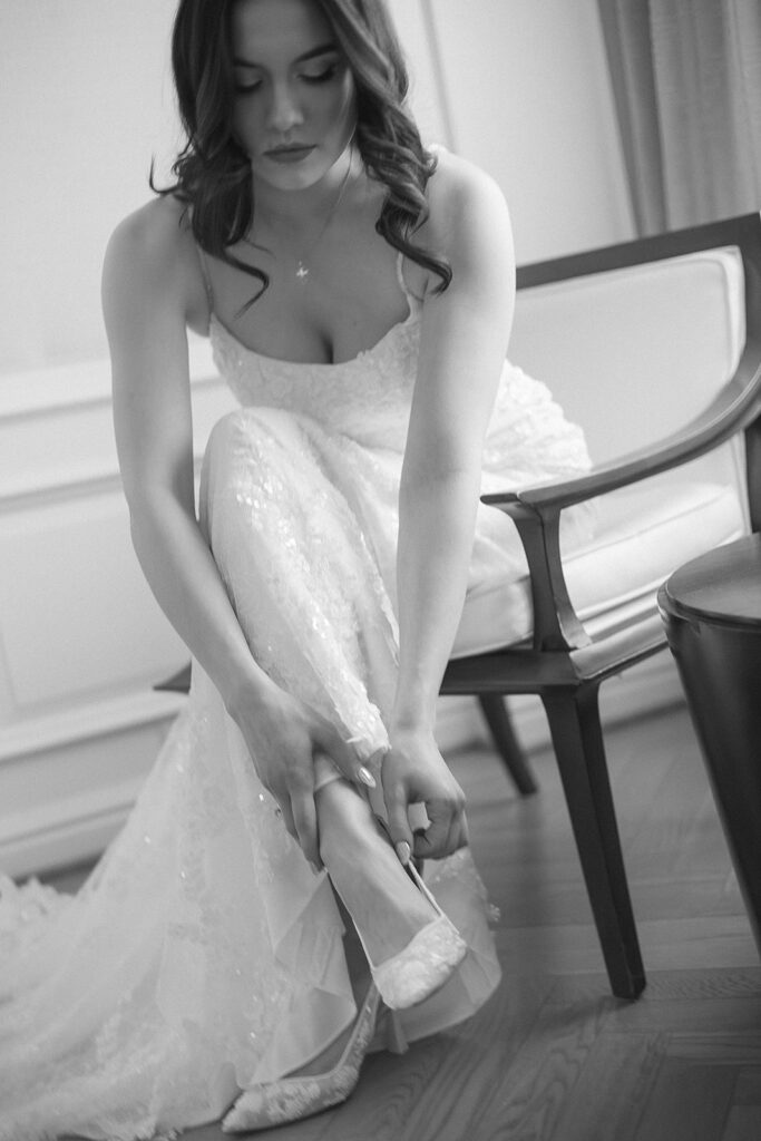 A woman in a wedding dress sits on a chair, leaning forward to put on a high-heeled shoe. The black-and-white photo captures an intimate, quiet moment before a wedding.