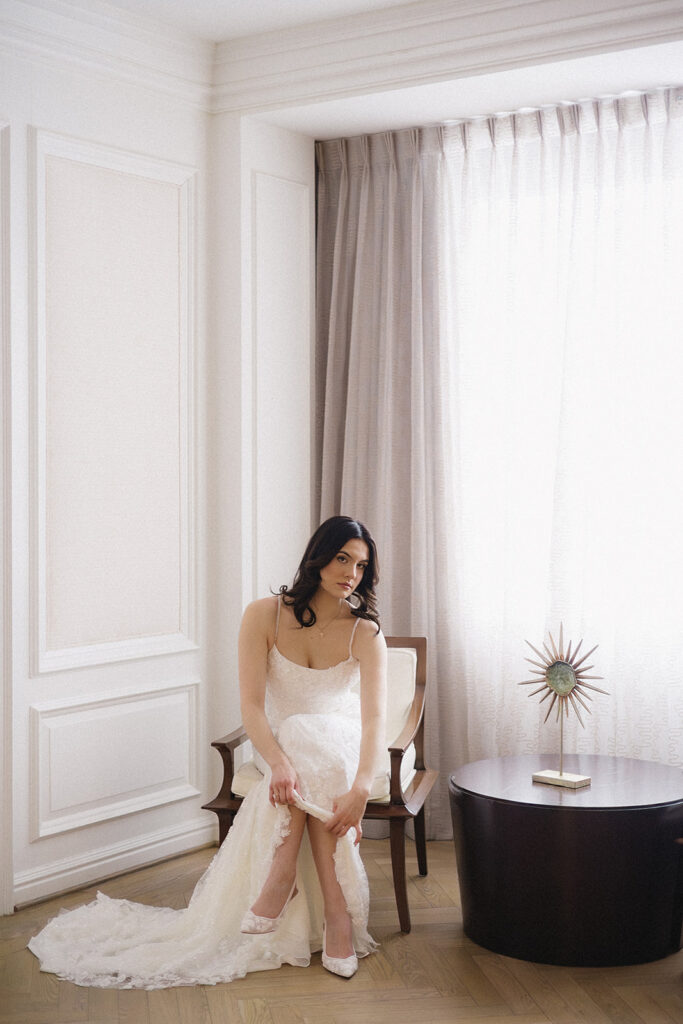 A bride in a white lace gown sits on a chair near a window, adjusting her shoe. Soft natural light fills the elegant, minimalist room with white paneled walls, sheer curtains, and a round dark table with a decorative piece.