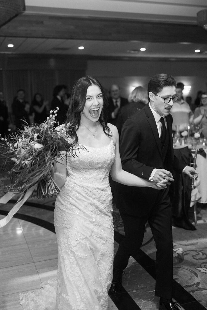 A joyful bride in a lace wedding dress holds a bouquet and smiles widely while walking hand in hand with her partner, who wears a suit, at a wedding reception with guests in the background.