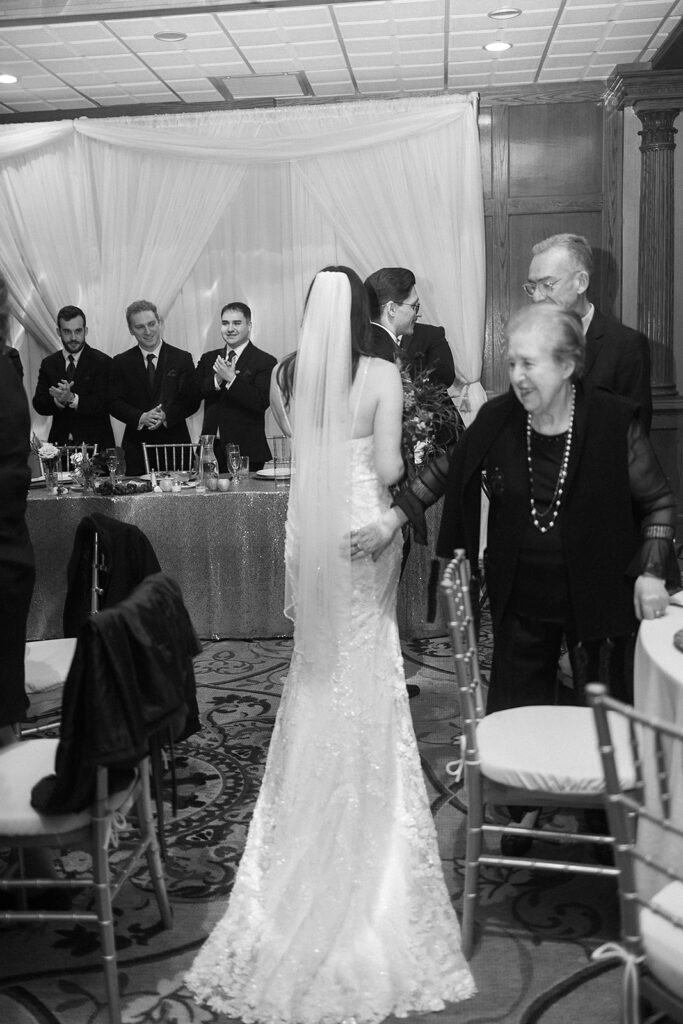 A bride in a lace gown and veil stands among guests at a wedding reception, facing a table where people in formal wear are standing and smiling. The atmosphere is celebratory and warm.
