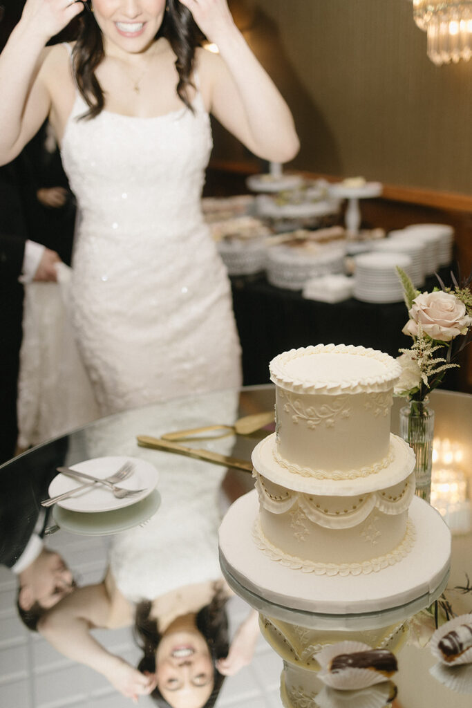 A smiling bride in a white dress stands behind a two-tiered decorated wedding cake on a glass table, which reflects her image. A plate with a fork and a small flower vase are beside the cake.