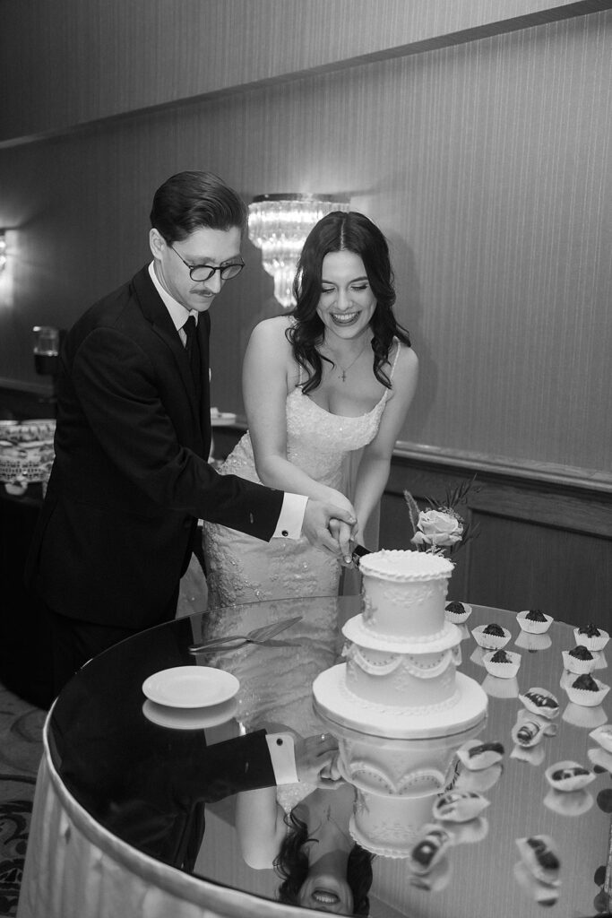 A bride and groom, both smiling, cut a wedding cake together at a decorated table. The scene is elegant, with cupcakes around the cake. The image is in black and white.