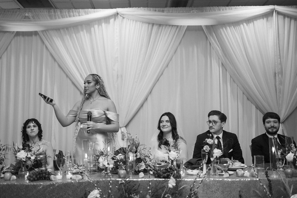 A woman in a light dress stands holding a microphone and a phone, speaking at a wedding reception table. Four guests, two women and two men, sit and listen. The table is decorated with flowers and draped fabric hangs behind them.