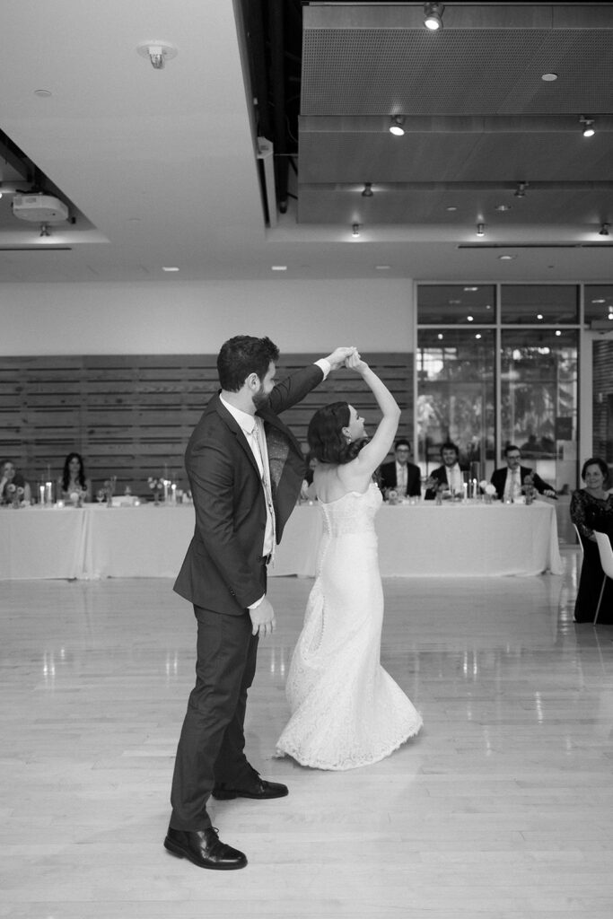 A groom in a suit twirls his bride, who is wearing a strapless wedding dress, as they dance together in a bright, modern reception hall with seated guests watching in the background.