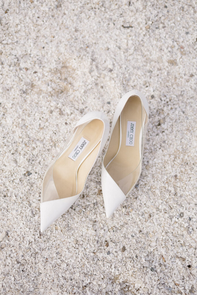 A pair of white pointed-toe Jimmy Choo high heels is placed side by side on a textured, light-colored, gravel-like surface.