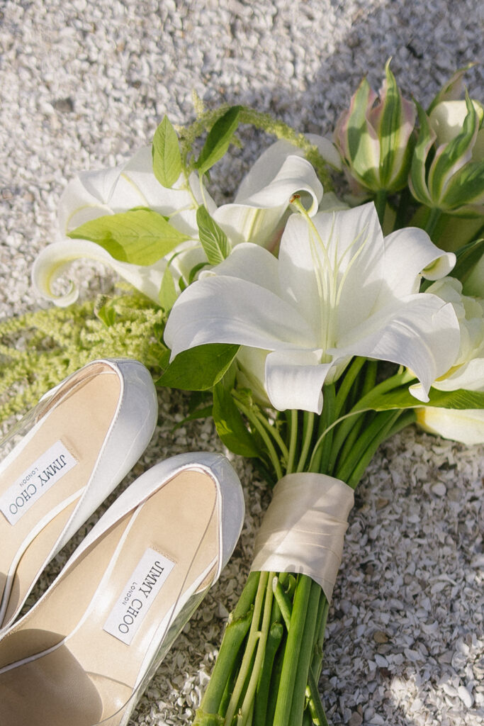 A bouquet of white lilies with green leaves lies on gravel next to a pair of white Jimmy Choo high-heeled shoes.