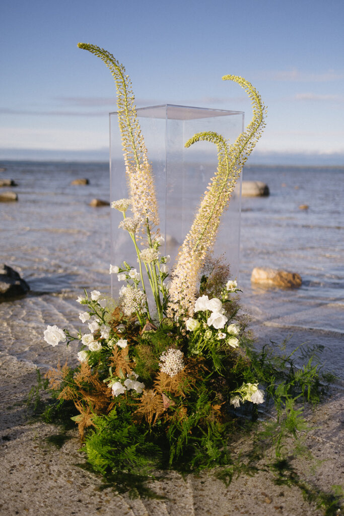 A modern floral arrangement with tall, spiky white flowers, lush greenery, and small white blooms, displayed in front of a clear box on a sandy beach with rocks and a calm sea in the background.