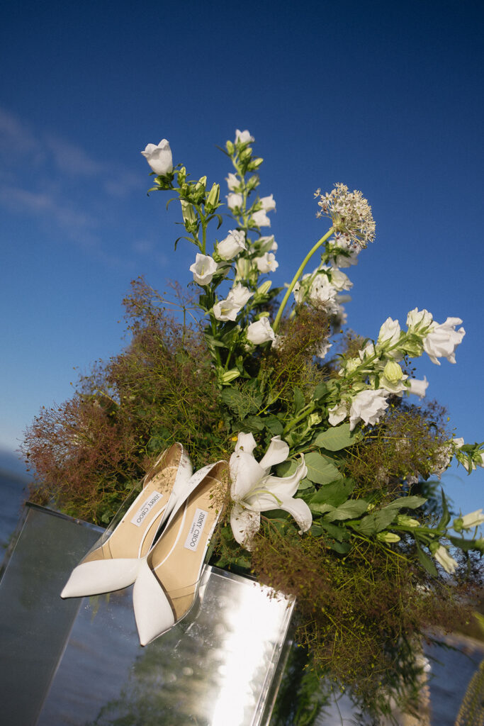 White high-heeled shoes are displayed on a reflective surface in front of a lush bouquet of white flowers and greenery, with a clear blue sky in the background.