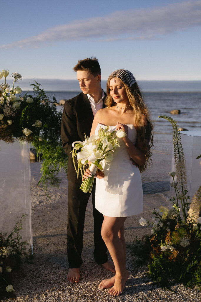 A barefoot couple stands on a beach at sunset, dressed in formal wedding attire. The woman holds a bouquet of white flowers and wears a white dress with a beaded headpiece; the man in a black suit stands behind her, surrounded by floral arrangements.