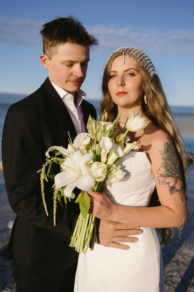 A bride in a white dress holding a bouquet of white flowers stands next to a groom in a black suit. They are outdoors with blue sky and water in the background. The bride has a floral tattoo on her arm and long hair.