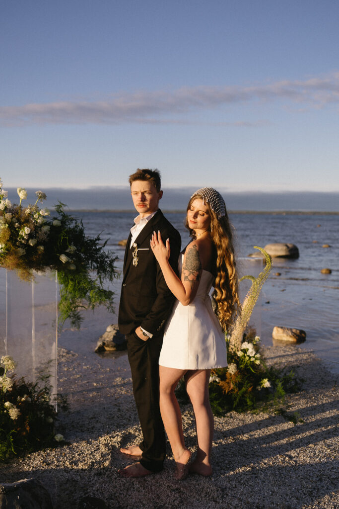A couple stands on a beach at sunset, dressed formally; the man in a black suit and the woman in a short white dress with a pearl headpiece. They are surrounded by floral arrangements and face the camera.