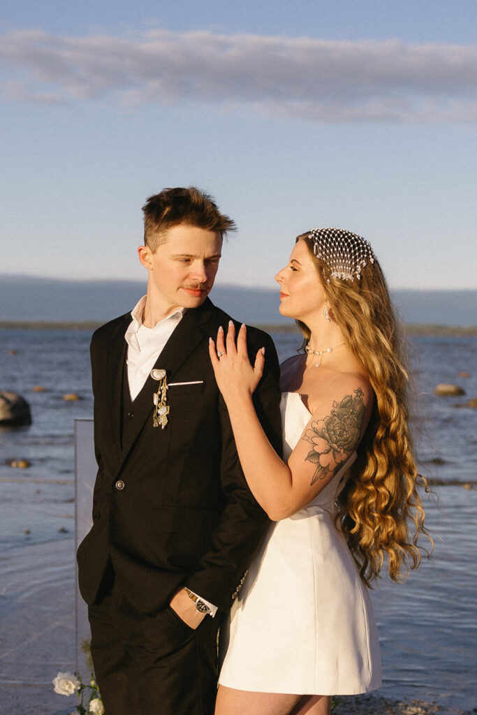 A couple stands by the water; the man wears a black suit and the woman, with long wavy hair and a pearl headpiece, wears a white dress and has a floral tattoo on her arm. They gaze at each other affectionately.