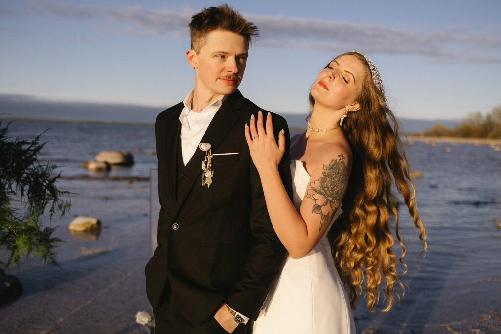 A bride and groom stand by the water at sunset. The groom wears a black suit and looks at the bride, who wears a white dress and tiara, with long wavy hair and a floral tattoo on her arm, resting her hand on his shoulder.