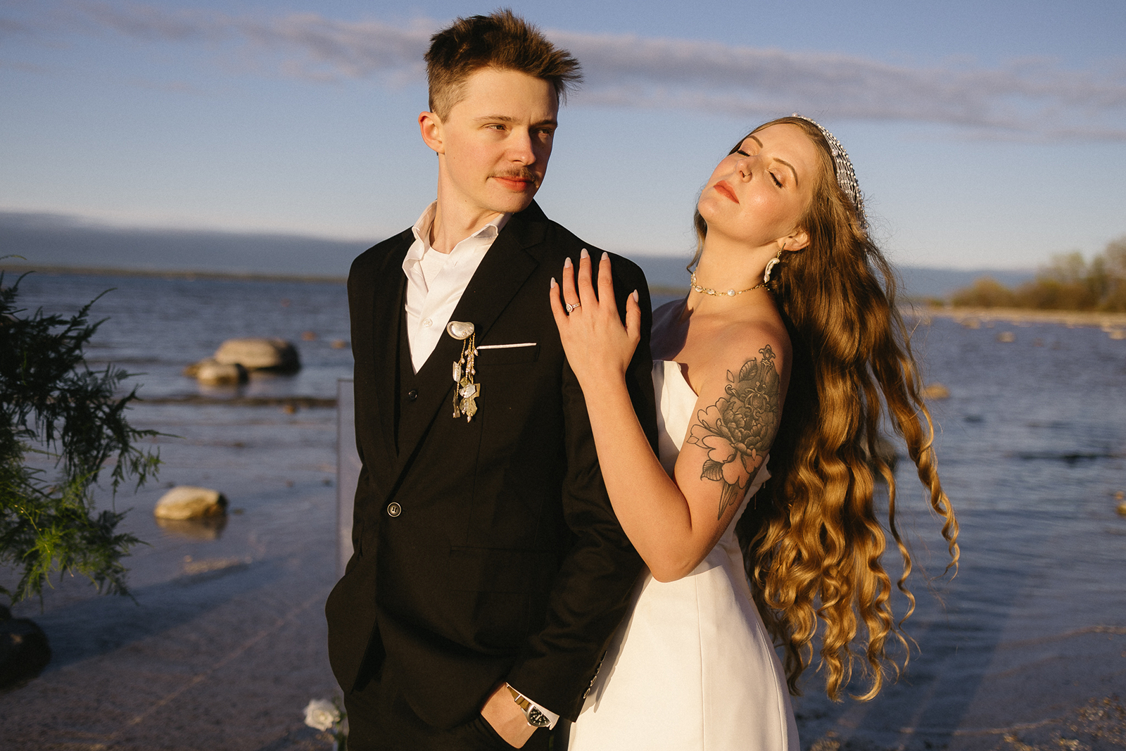 A bride and groom stand by the water at sunset. The groom wears a black suit and looks at the bride, who wears a white dress and tiara, with long wavy hair and a floral tattoo on her arm, resting her hand on his shoulder.
