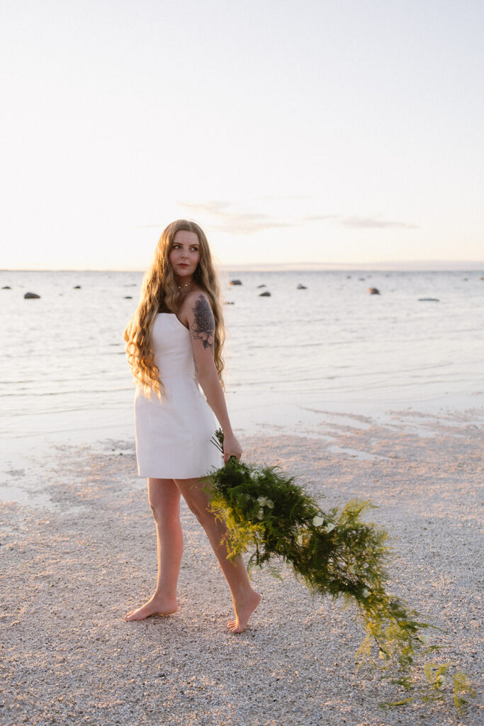 A woman with long hair in a white dress stands barefoot on a beach at sunset, holding a bundle of green foliage. The sea and scattered rocks are visible in the background under a clear sky.