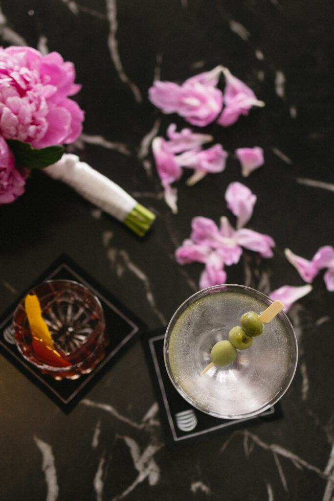 A martini with three green olives and a cocktail with an orange peel sit on black coasters on a dark marble surface, next to a bouquet of pink flowers and scattered pink petals.