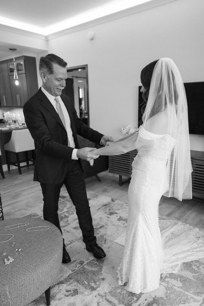 A man in a suit smiles as he holds hands with a bride in a white gown and veil. They stand in a modern living room, sharing a joyful moment before the wedding ceremony.