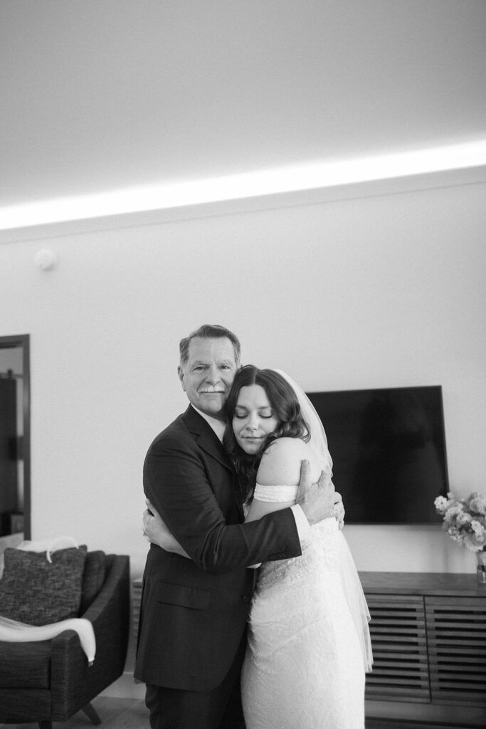 A bride in a wedding dress hugs an older man in a suit, both smiling softly. They stand indoors near a TV, flowers on a cabinet, and a sofa in the background. The moment appears emotional and heartfelt.