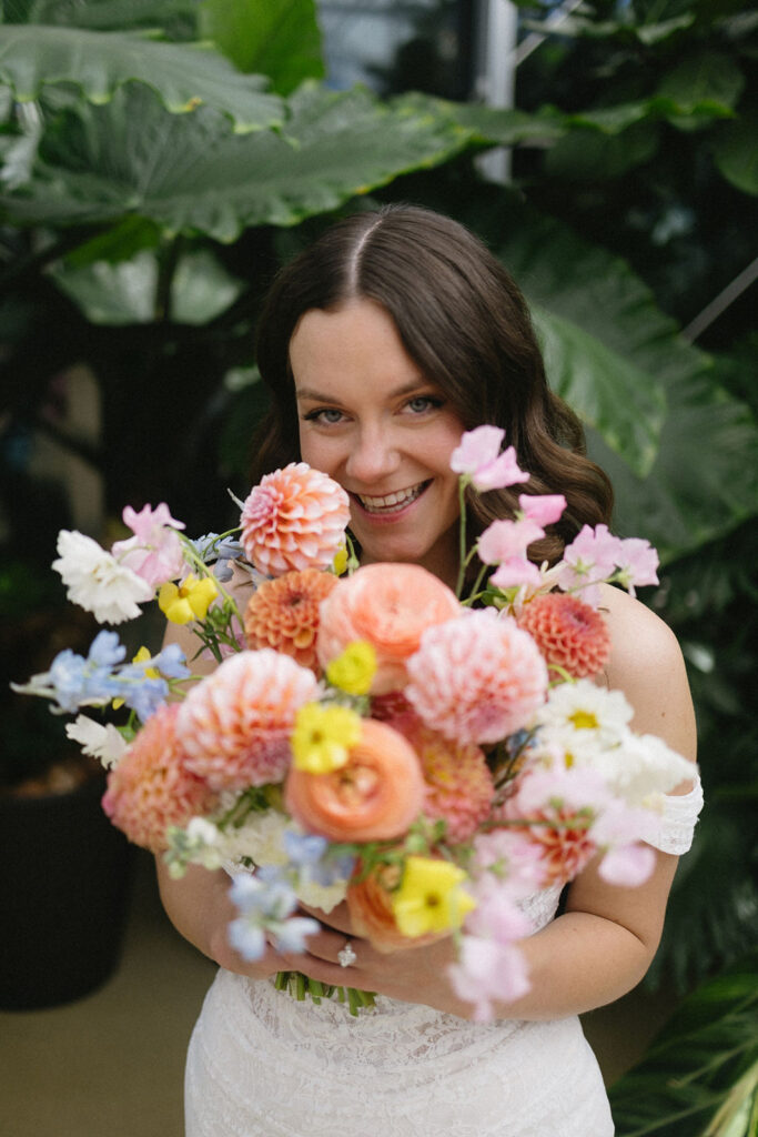 A woman in a white dress smiles and holds a colorful bouquet of flowers in front of lush green foliage.