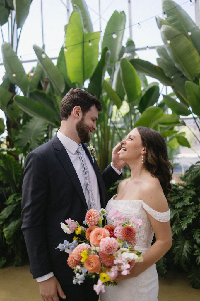 A bride in an off-shoulder white dress holds a colorful bouquet and smiles up at the groom in a dark suit, who gazes at her lovingly. They stand in a lush, indoor garden with large green tropical plants.