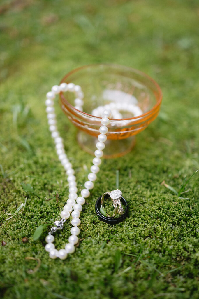 A pearl necklace and two rings, including a diamond engagement ring, rest on green moss next to a small orange glass bowl.