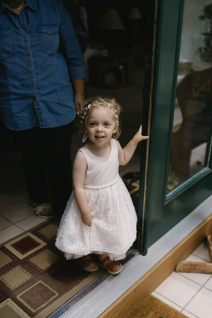 A young girl in a white dress stands smiling in a doorway, holding the door with one hand. She has curly hair with a flower headband. An adult stands behind her, partially visible, wearing a blue shirt.