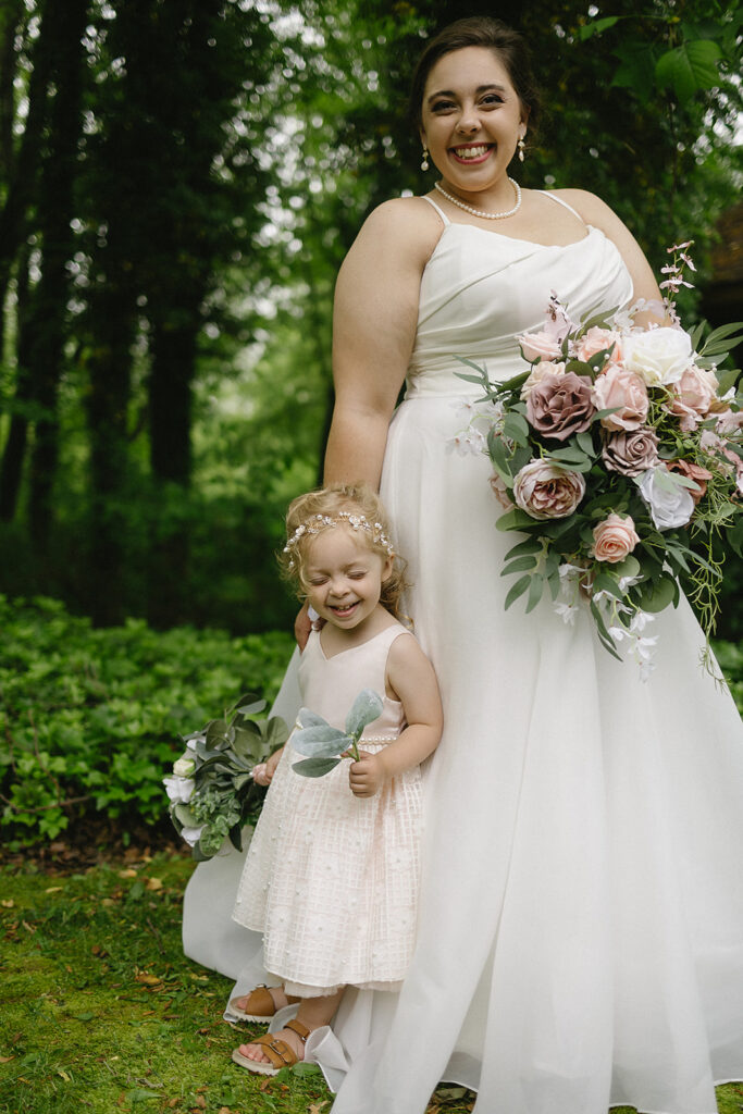 A smiling bride in a white gown and pearl necklace stands outdoors holding a large bouquet of pastel flowers, next to a happy little girl in a light dress and floral headband, both surrounded by greenery.