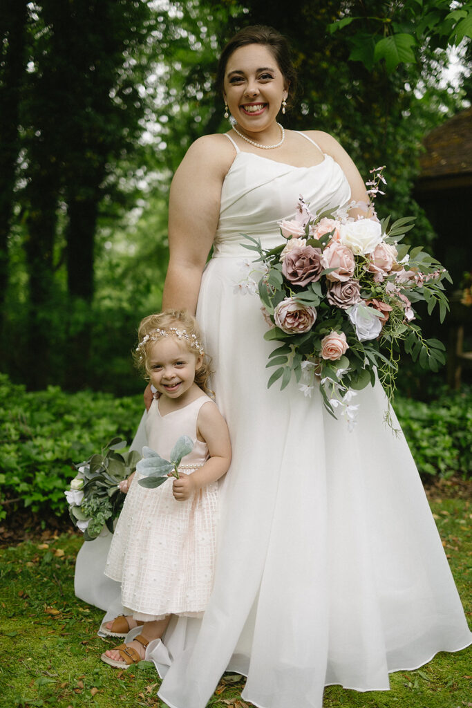 A smiling bride in a white dress holds a large bouquet of pink and white flowers, standing beside a happy young flower girl in a light pink dress, surrounded by greenery outdoors.