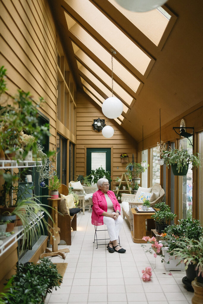 An older woman in a bright pink jacket sits on a chair in a sunlit indoor garden room with wooden walls, many green plants, white tile floor, and skylights above.